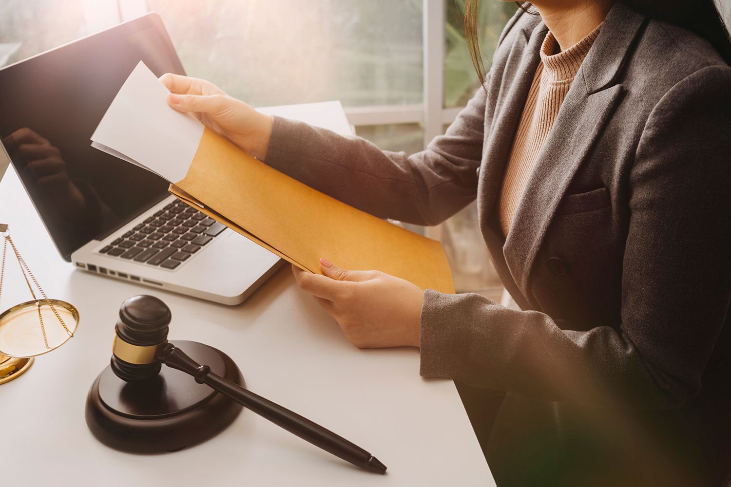 Person in suit opening an envelope, with a laptop, gavel, and scale on a desk.