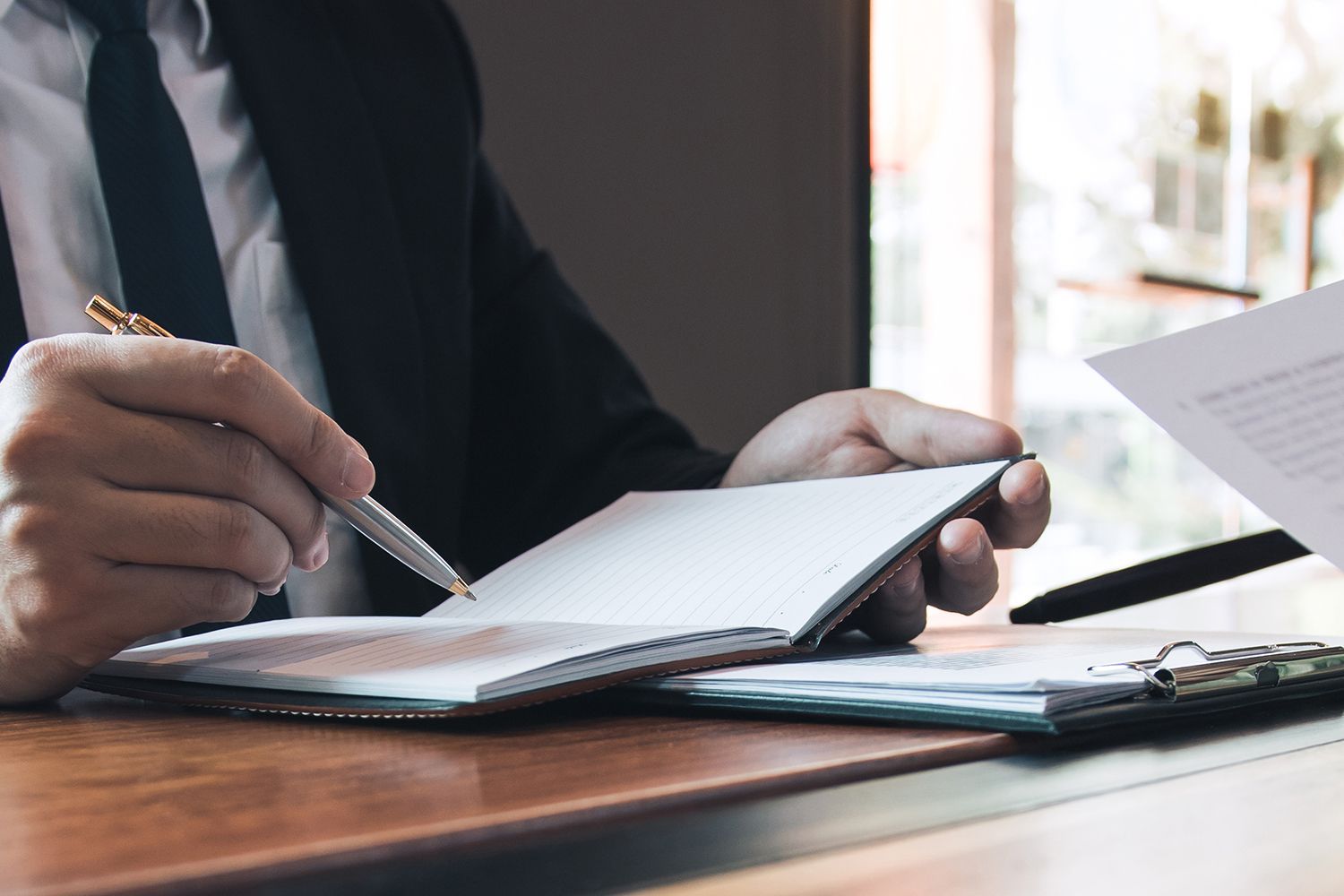 Person in suit writing in notebook with a pen at a desk.