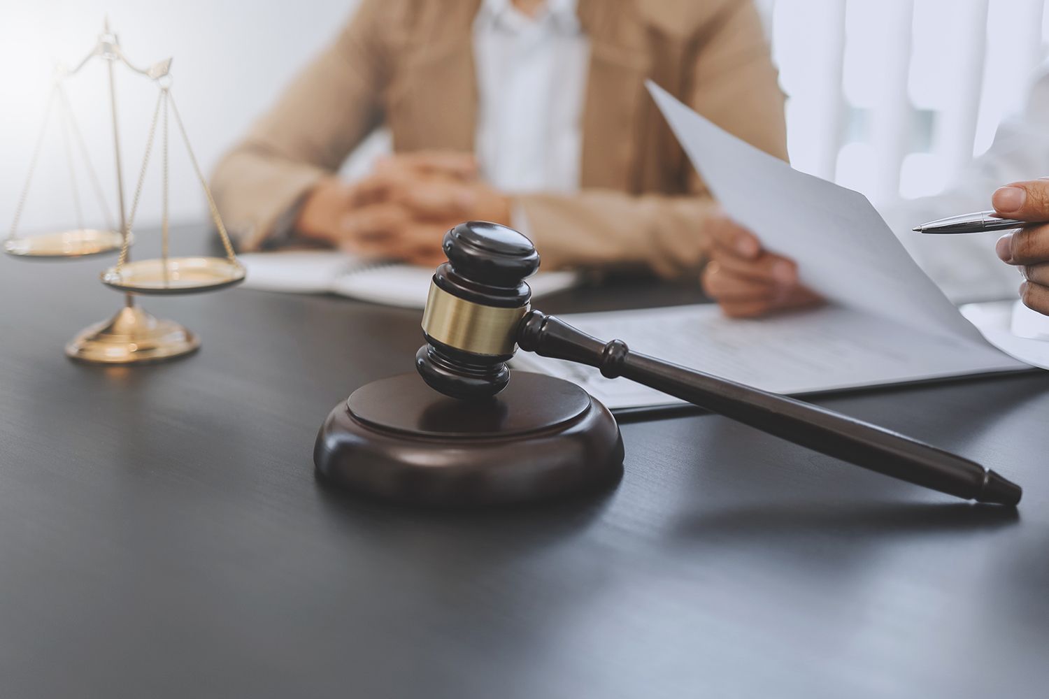 Gavel on a table with scales of justice and people reviewing documents. Gavel on a table with scales of justice and people reviewing documents.