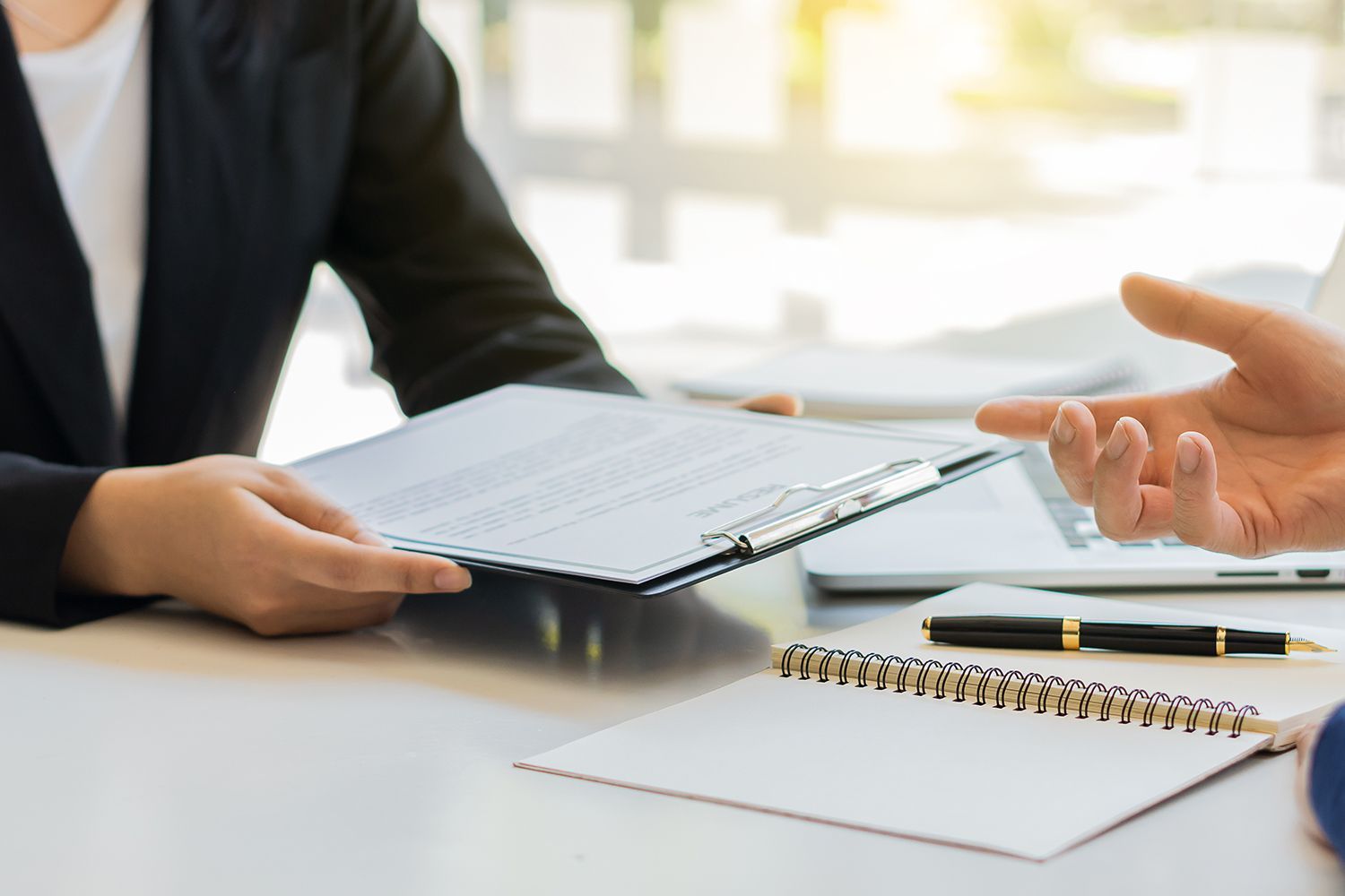 Person in suit handing document to another person, office setting with laptop and notepad. Person in suit handing document to another person, office setting with laptop and notepad.