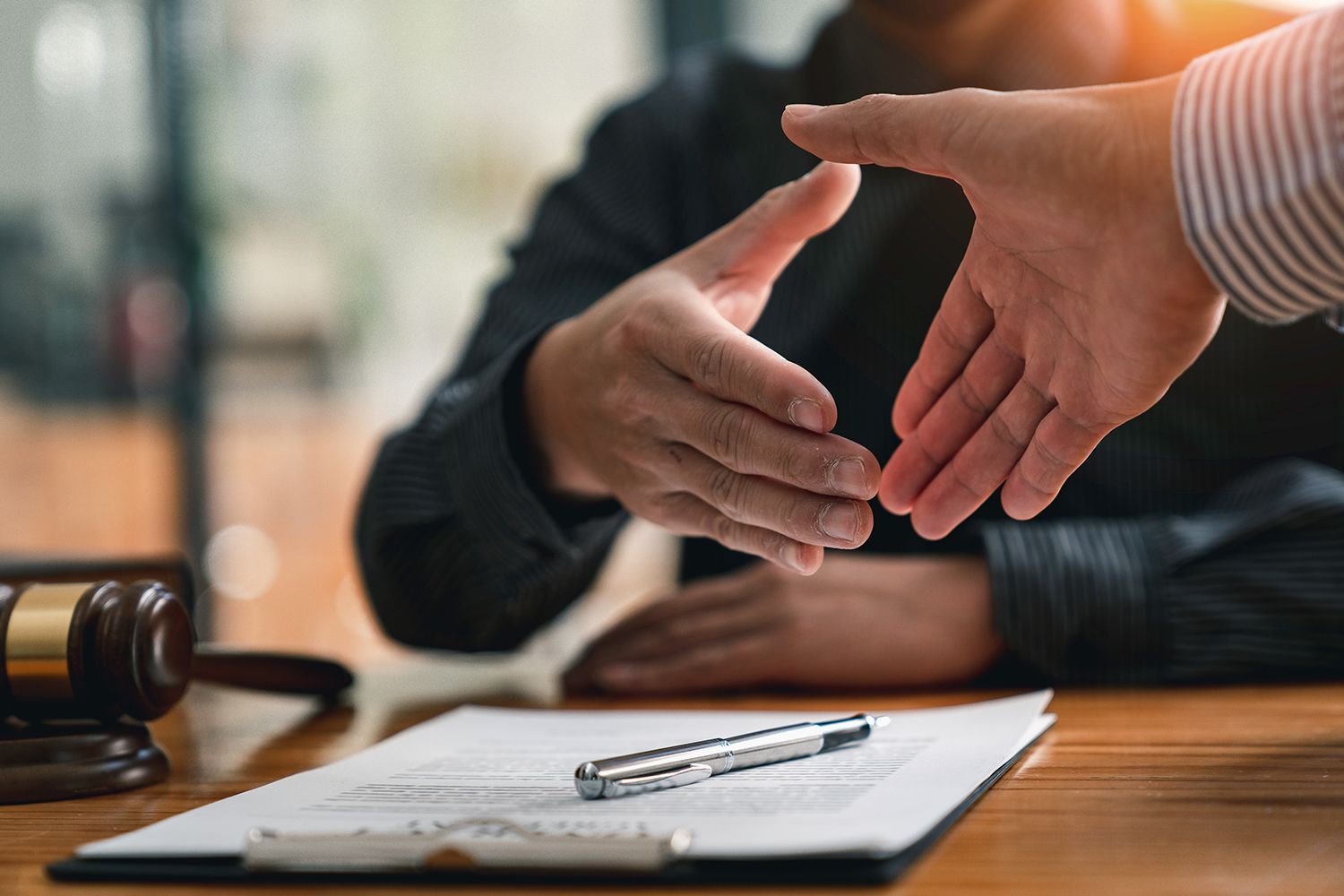 Two hands reaching for a handshake over a contract on a table with a gavel nearby.