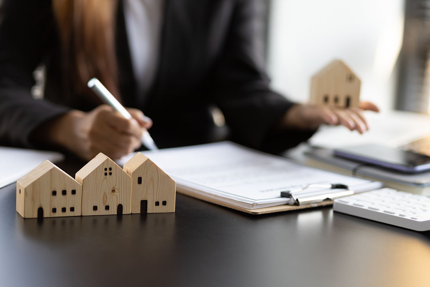 Person writing on documents with miniature wooden houses and calculator on a desk.