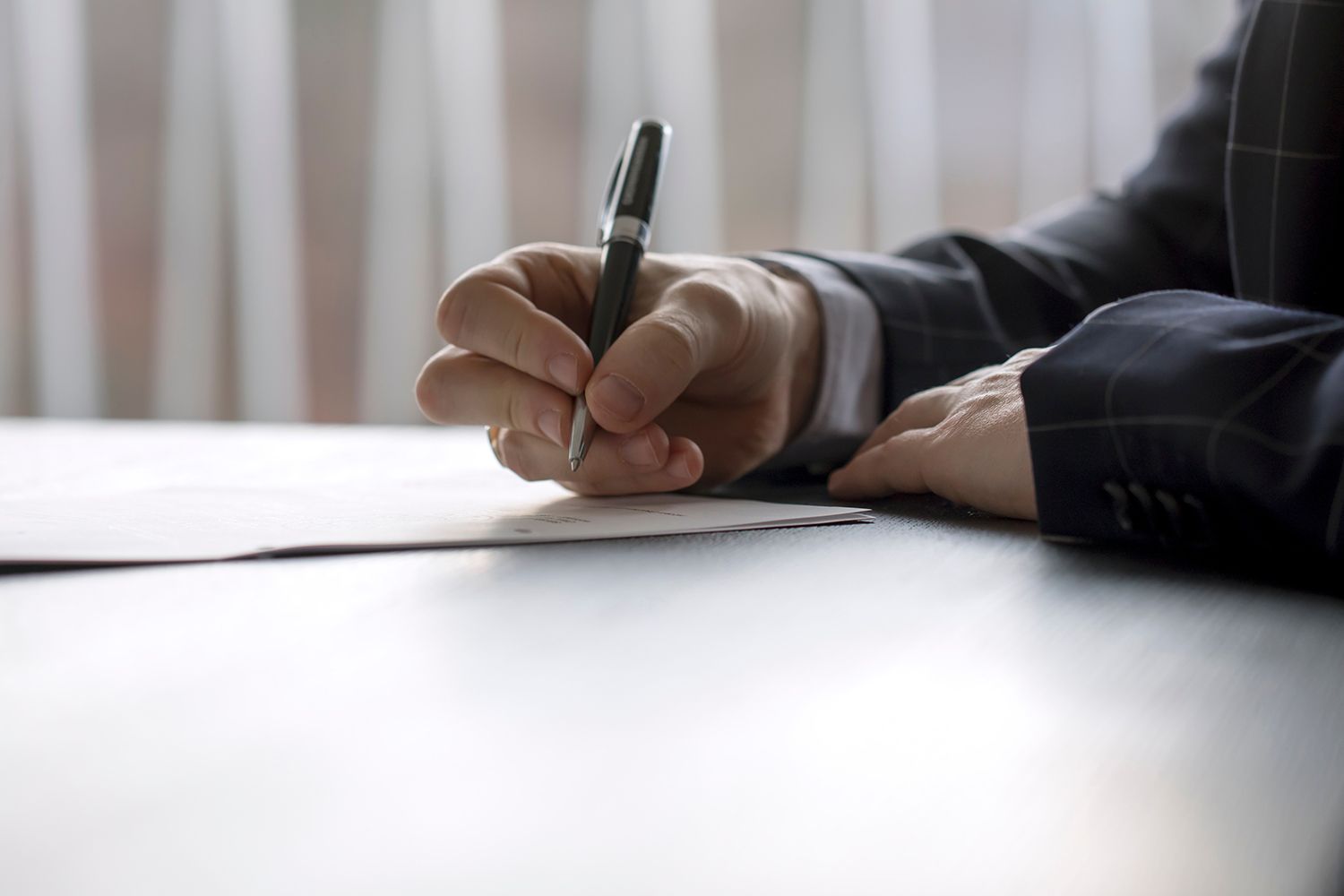 Person in suit writing with pen on a white paper at a table.