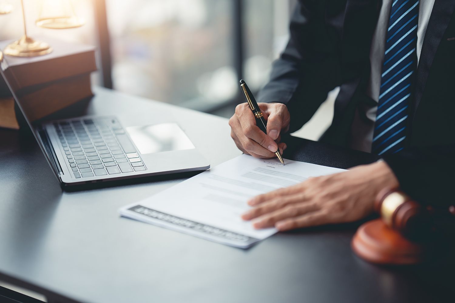 Person in suit signing document at desk with laptop, gavel, and books. Person in suit signing document at desk with laptop, gavel, and books.