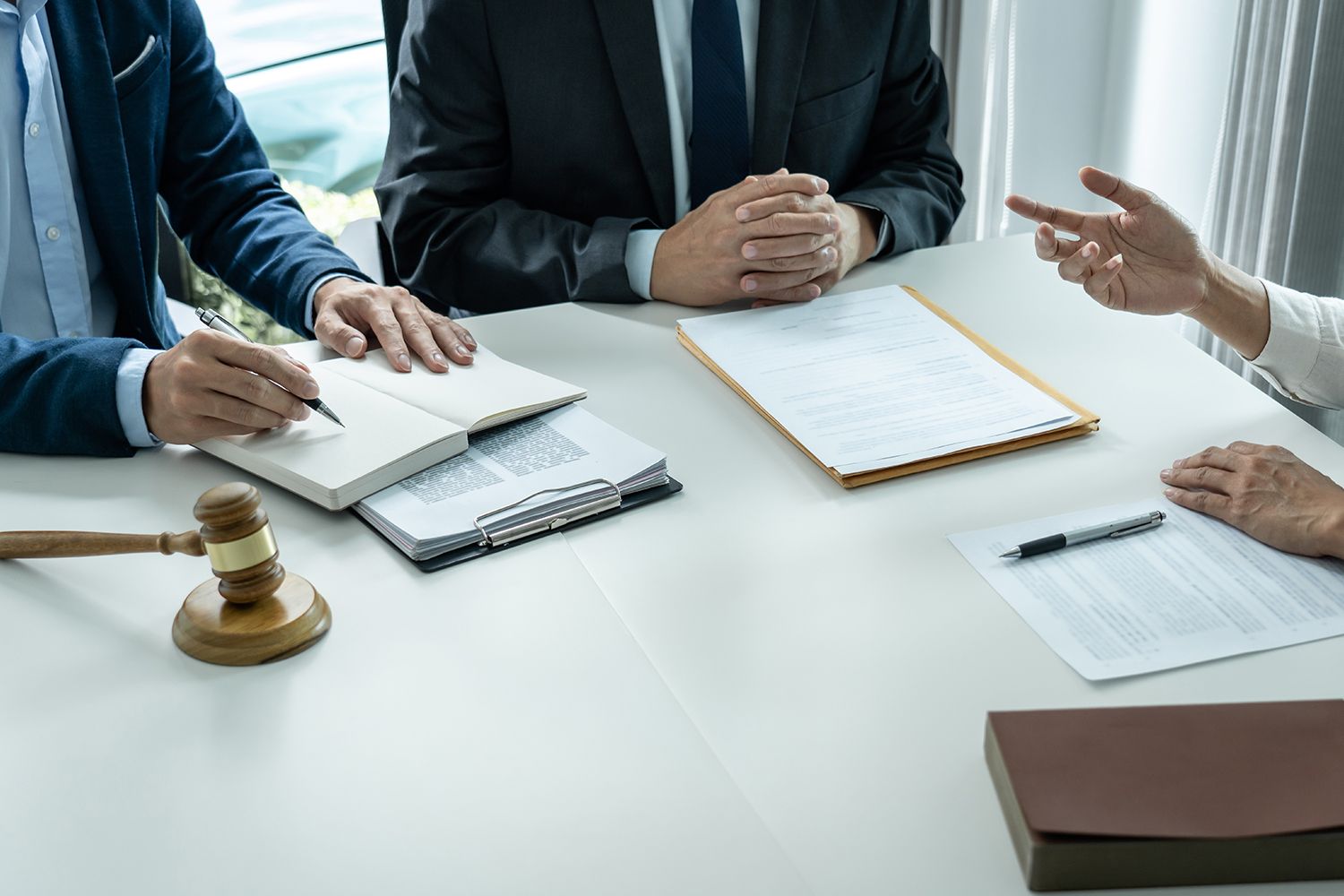 Three people in suits at a table with documents, a gavel, and a notepad. Someone gestures.