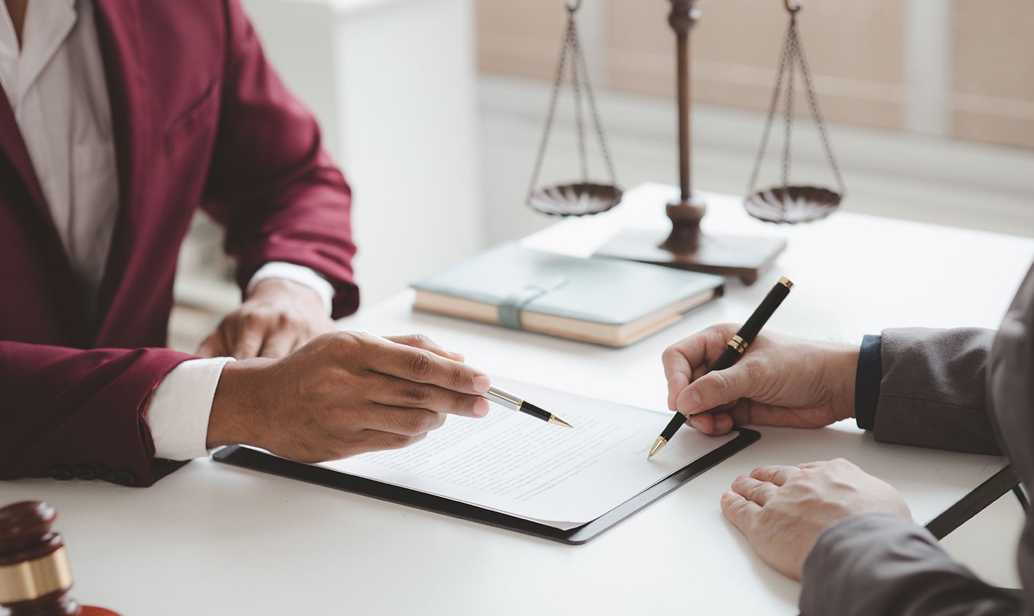 Two people at a desk, reviewing document; one points, other writes. Scales of justice in background.