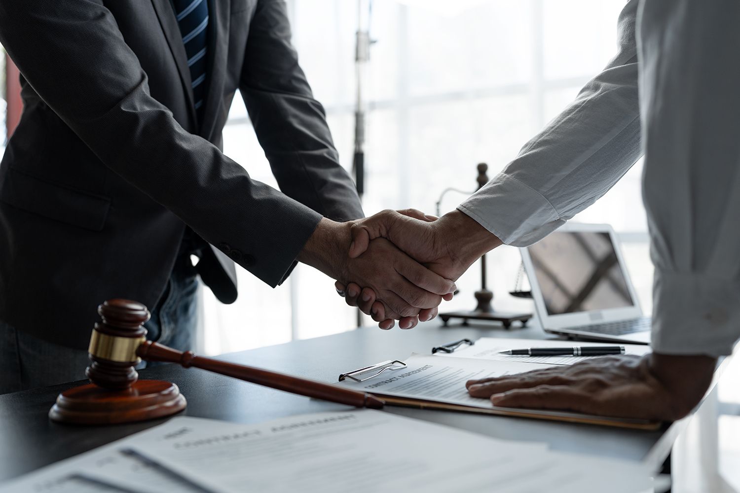 Two people shaking hands over a contract on a desk, gavel and scales of justice in background.