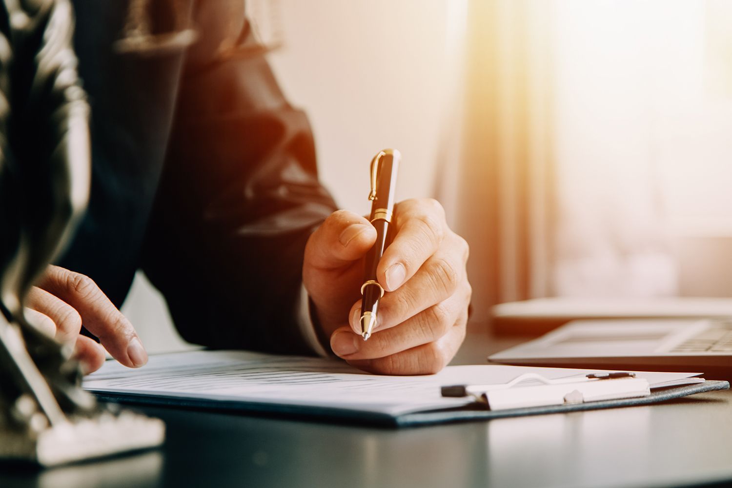 Person in suit signing documents with a pen at a desk, sunlight in background.