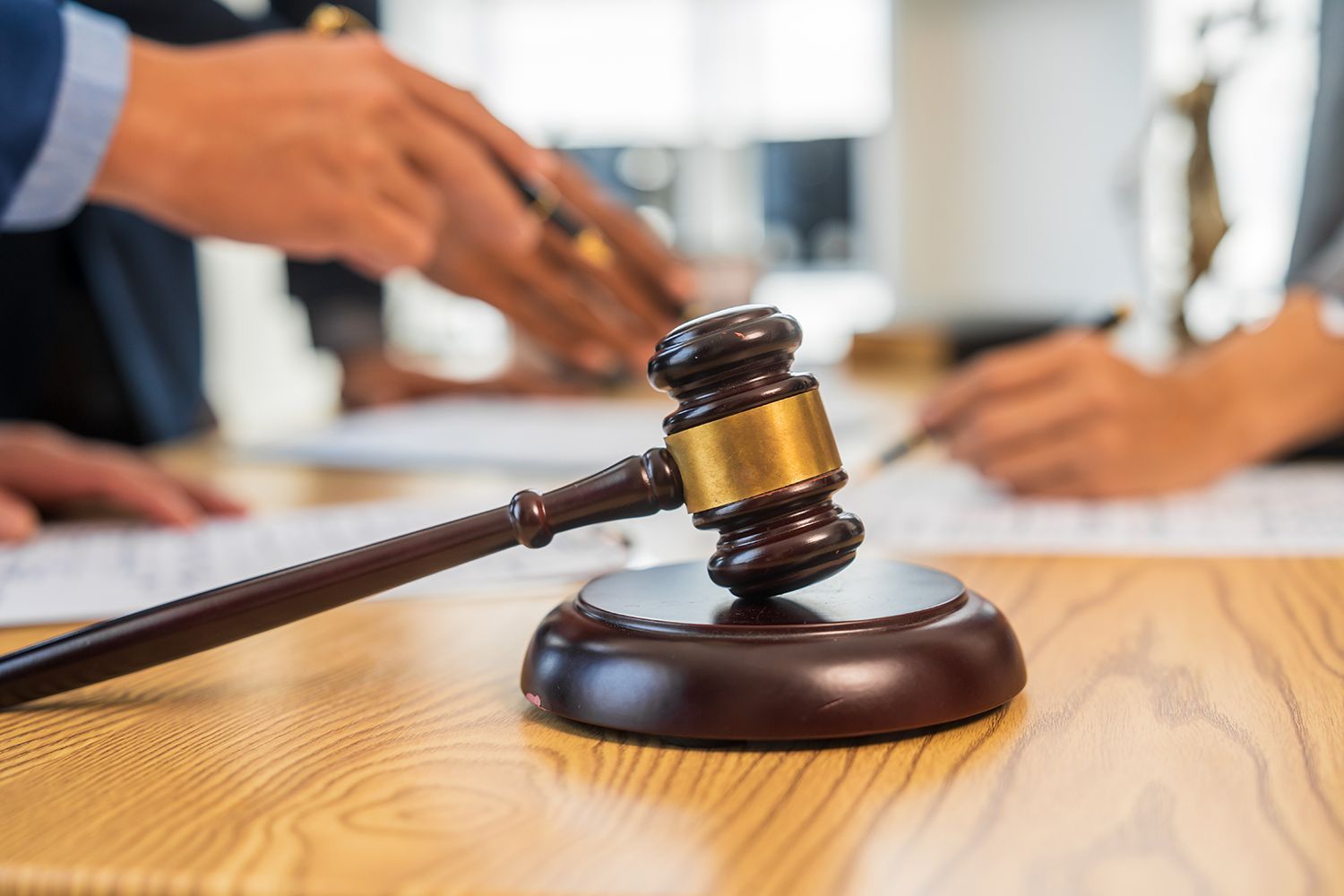 Gavel on wooden table with people blurred in the background, possibly in a courtroom.