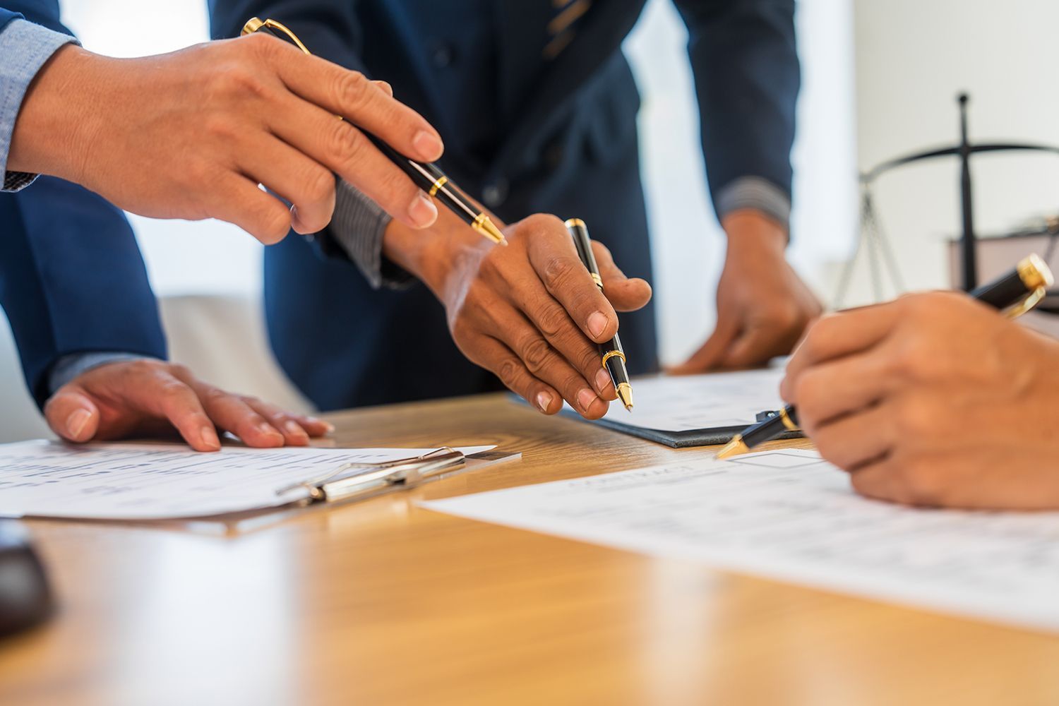 Hands pointing at documents on a wooden table. Three people wearing suits, signing a contract.