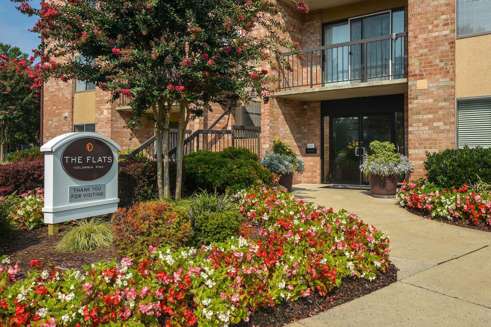 Brick apartment building entrance with glass doors and colorful flower beds along a walkway.