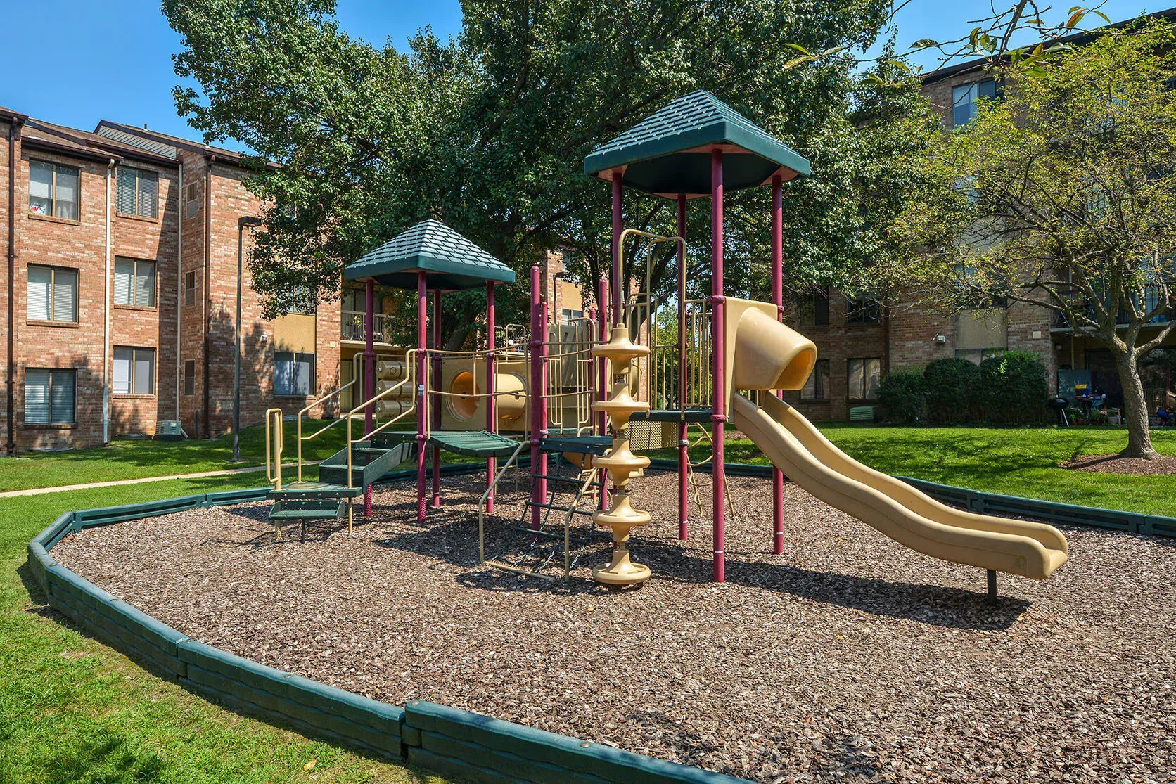 Playground with slides, climbing structures, and protective border in front of a brick apartment building.