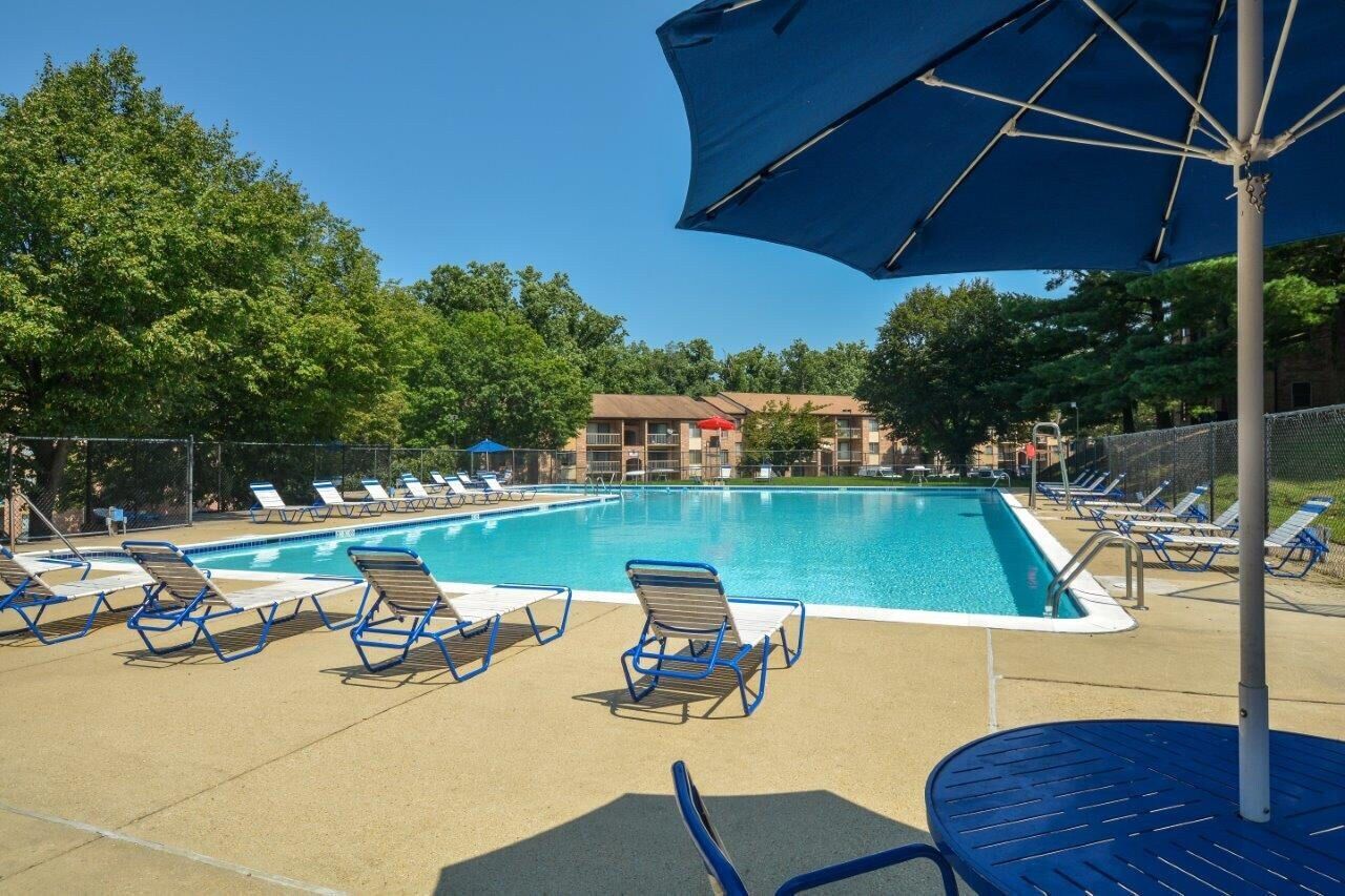 Swimming pool with blue umbrella, lounge chairs, and trees on a sunny day.