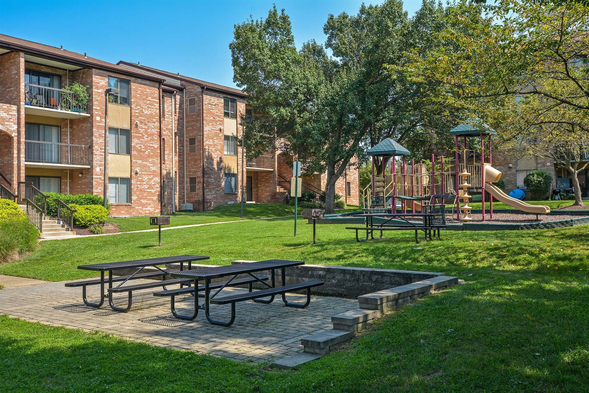Courtyard with brick apartment buildings, a playground, and picnic tables on a grassy lawn.