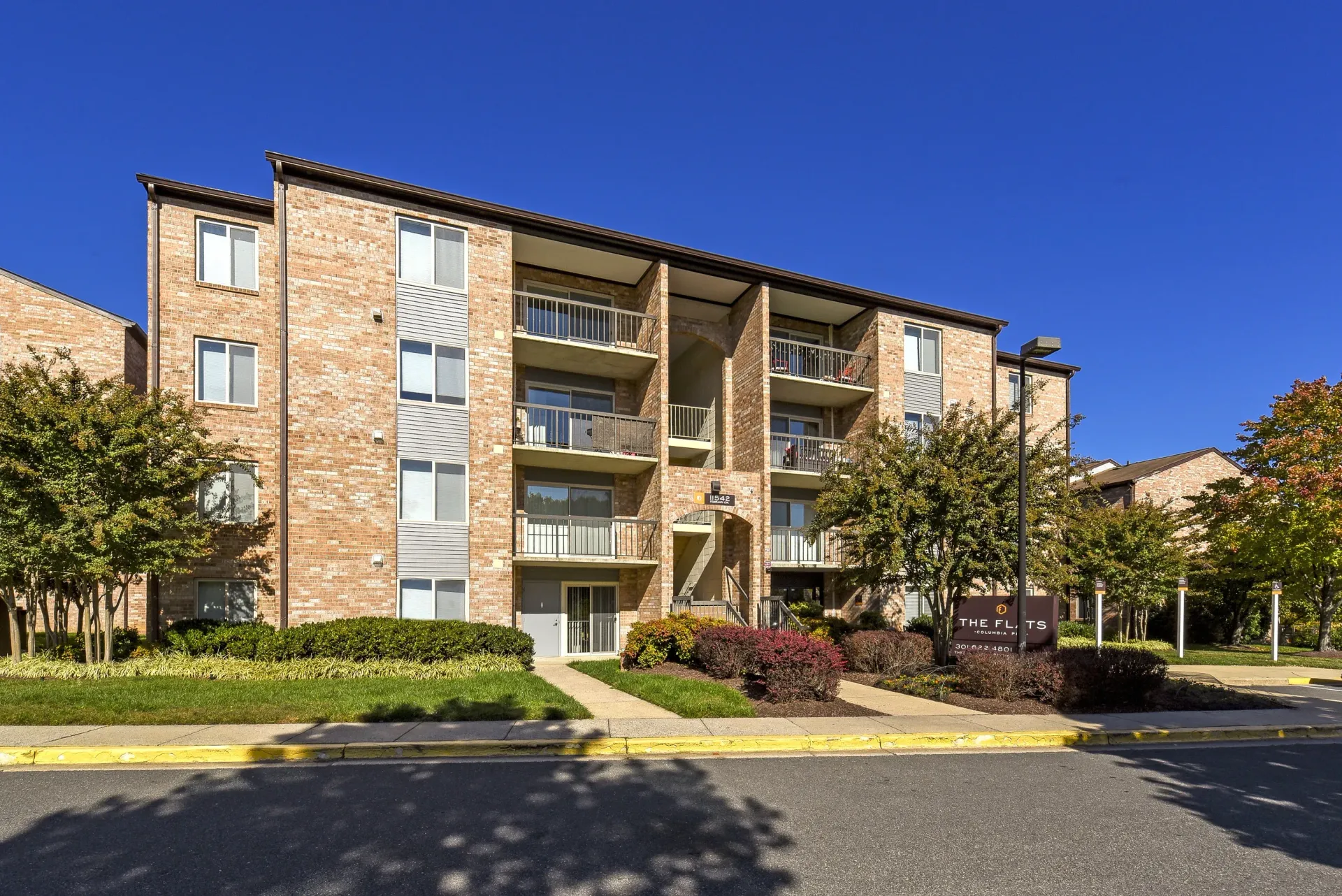 Exterior view of a brick apartment building with balconies and landscaped entry under a blue sky.
