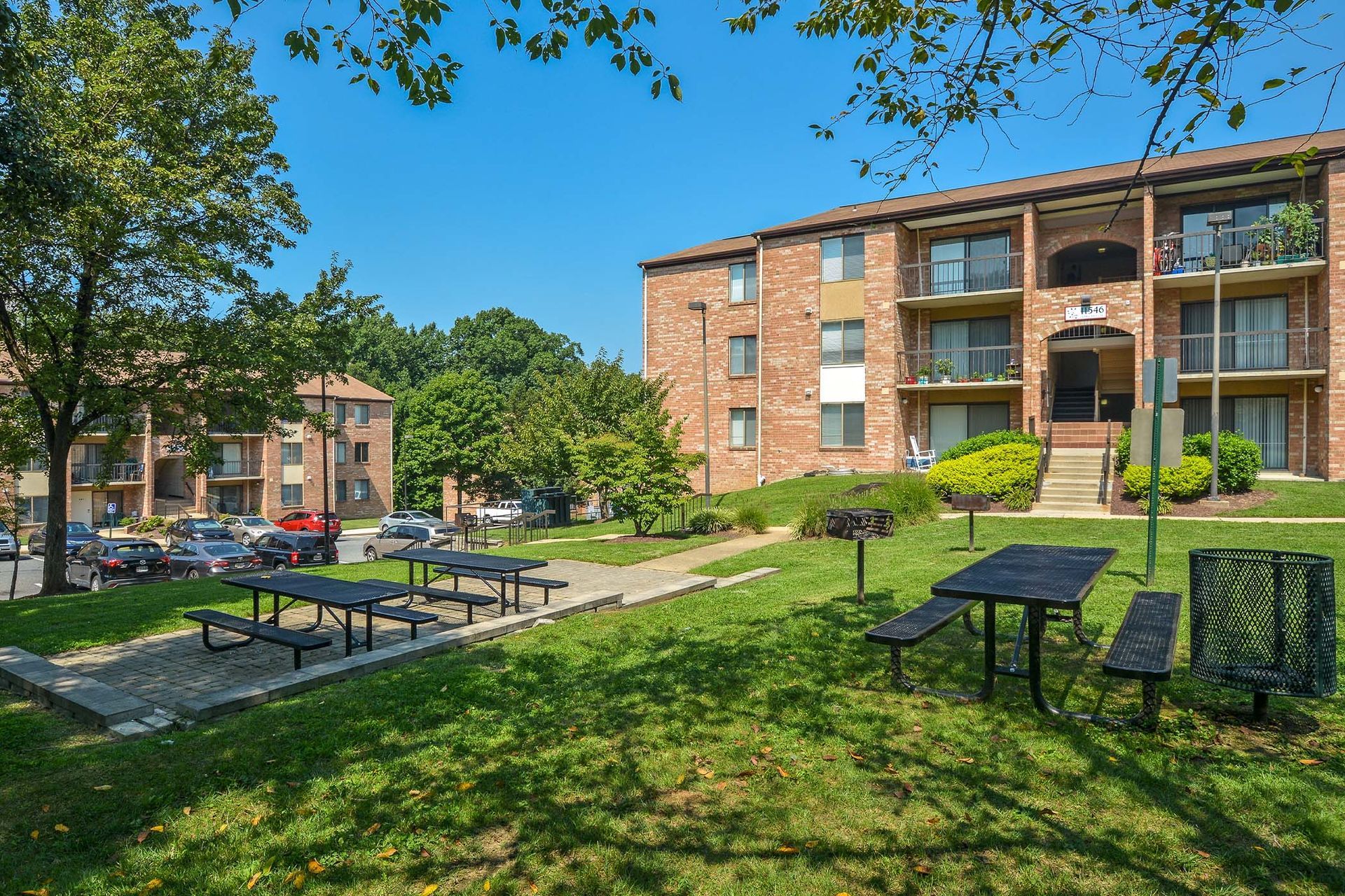Exterior view of brick apartment buildings with a grassy courtyard, trees, and picnic tables.