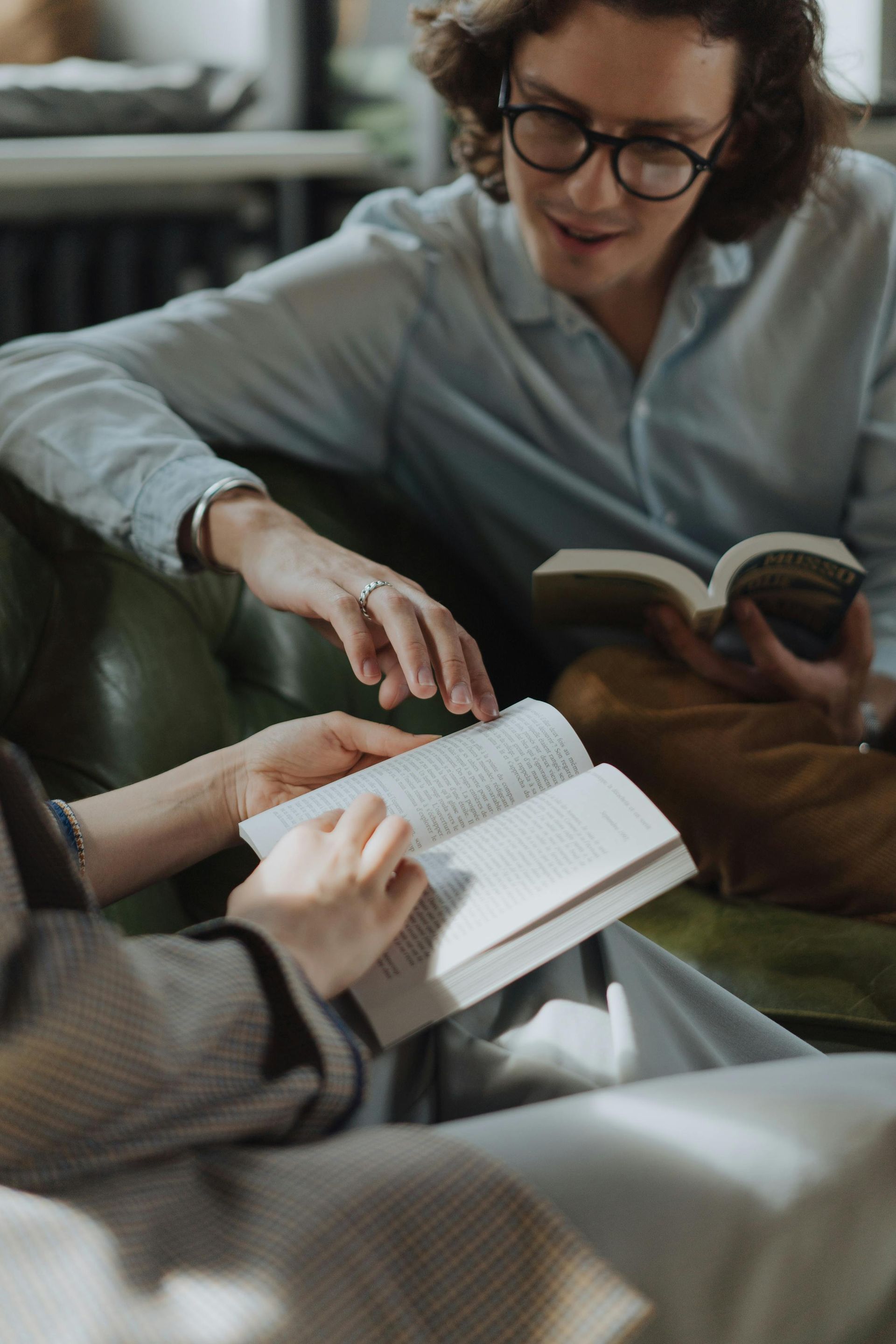 Two people seated, reading books. One points to a page, other smiles. Indoor setting.