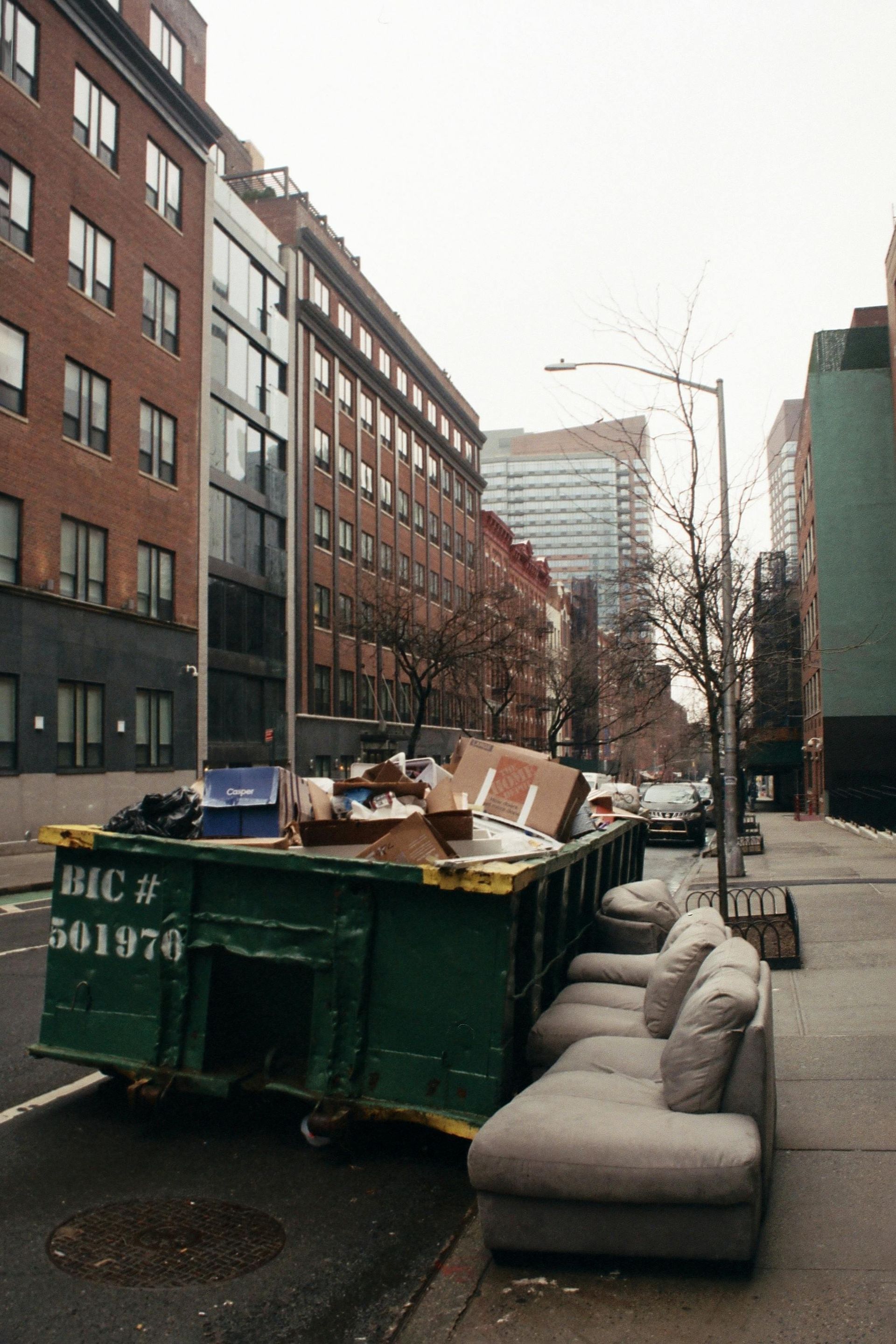 Green dumpster on city street; overflowing with trash, beside a light grey sofa. Brick buildings in background.