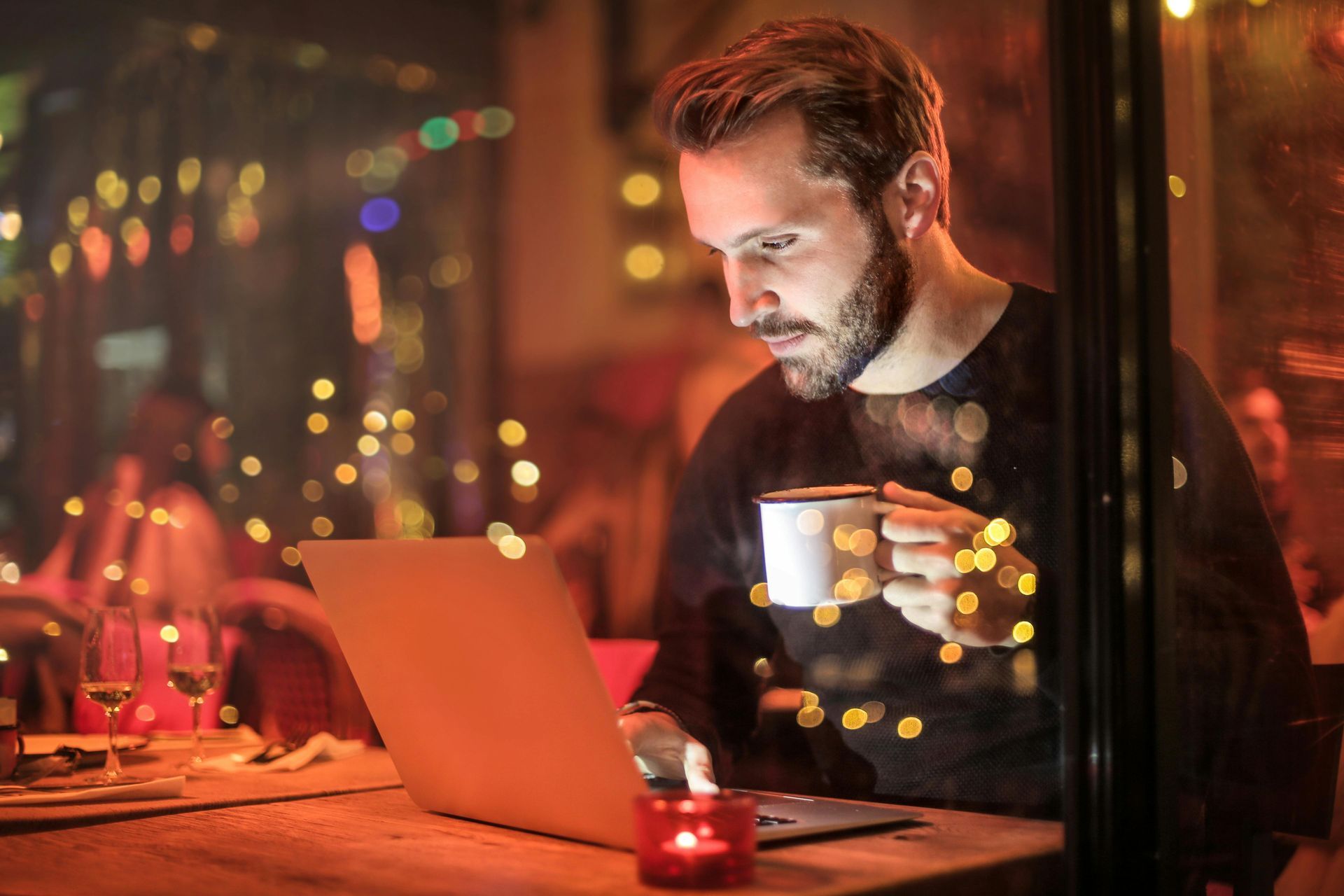 Man in a dimly lit cafe with laptop, holding a mug, looking intently at screen. Candle and string lights visible.