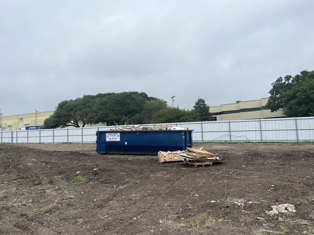 A blue dumpster in a fenced-off lot, next to piles of wood, under a cloudy sky.