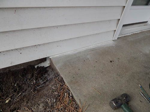 White siding meets a concrete patio. A gap reveals exposed dirt