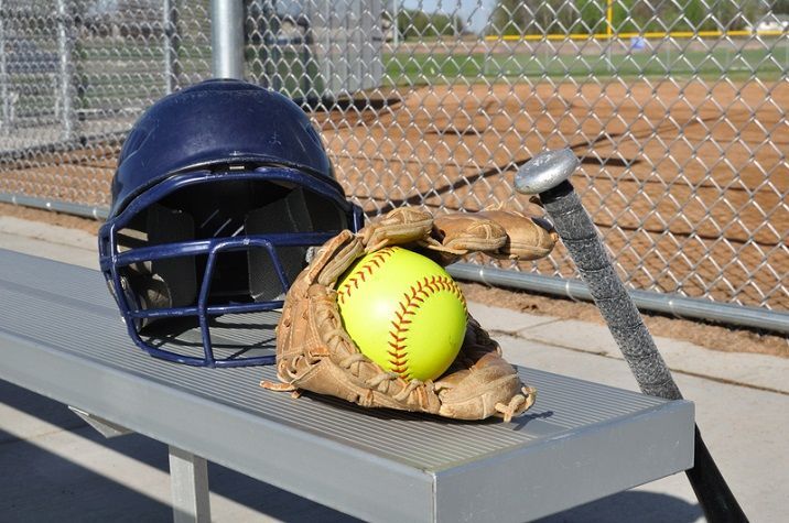A blue batting helmet, a leather glove holding a yellow softball, and a bat on a metal bench at a baseball field.