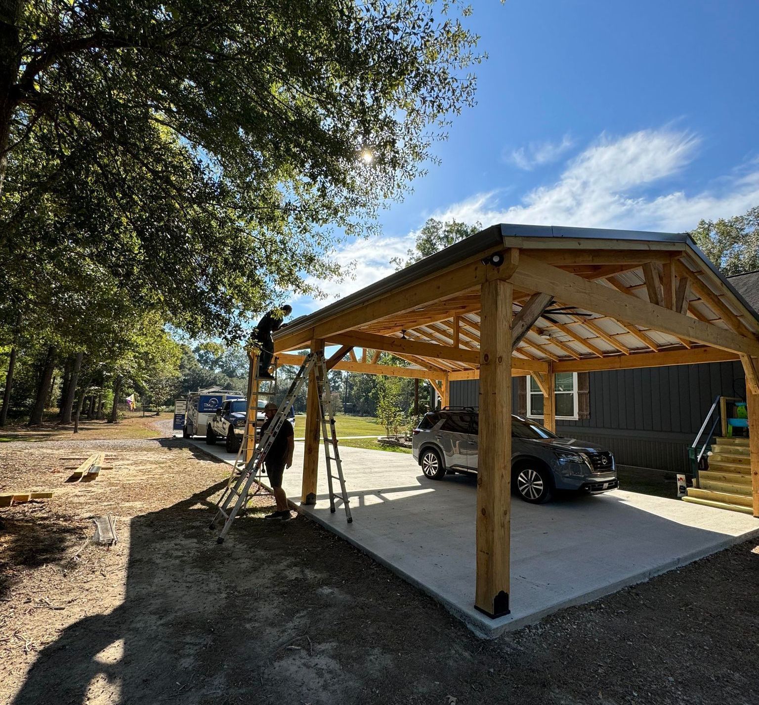 A car is parked under a wooden structure in a driveway.