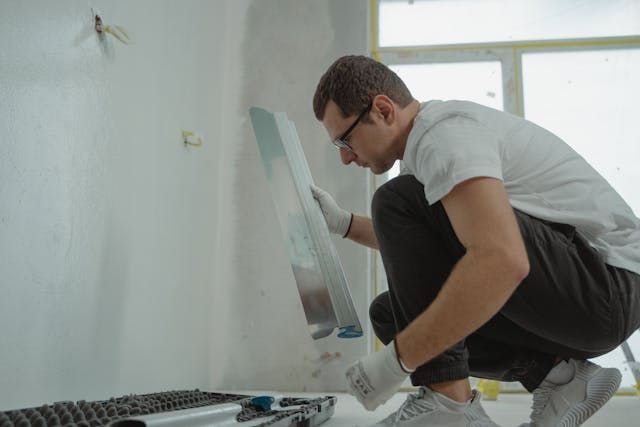 Person crouching down holding painting tools while wearing glasses