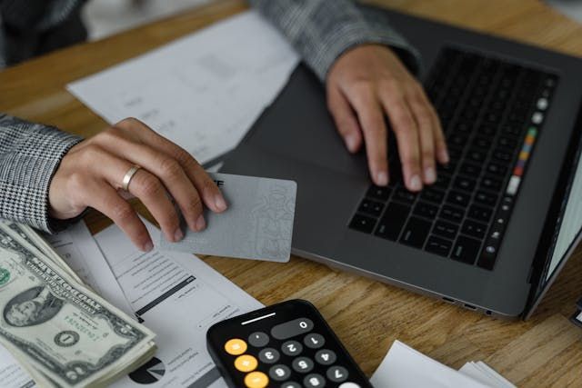A person is using a laptop while holding a bank card, there's money and a calculator sitting beside the laptop