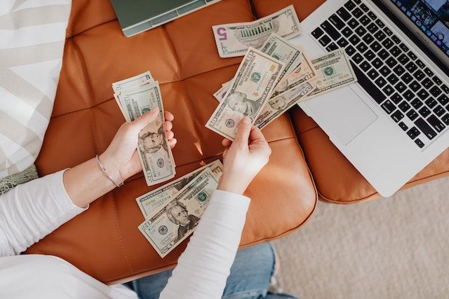 someone counting money on a sofa next to an open laptop