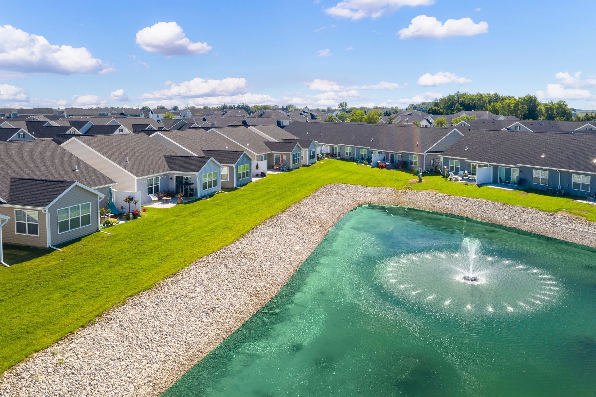Aerial view of houses along a pond with a fountain. Green grass and blue sky.