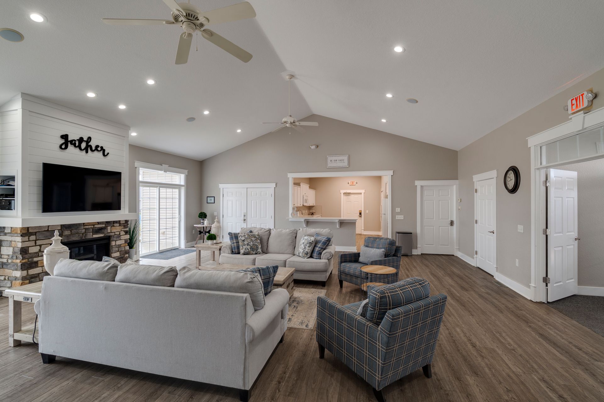 Spacious living room with sofas, fireplace, and open kitchen; light gray walls, wooden floor, and overhead fans.