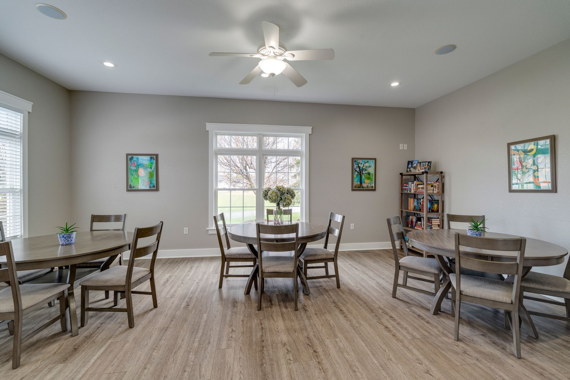 Dining area with round tables, chairs, and artwork. Window in the background and a ceiling fan.