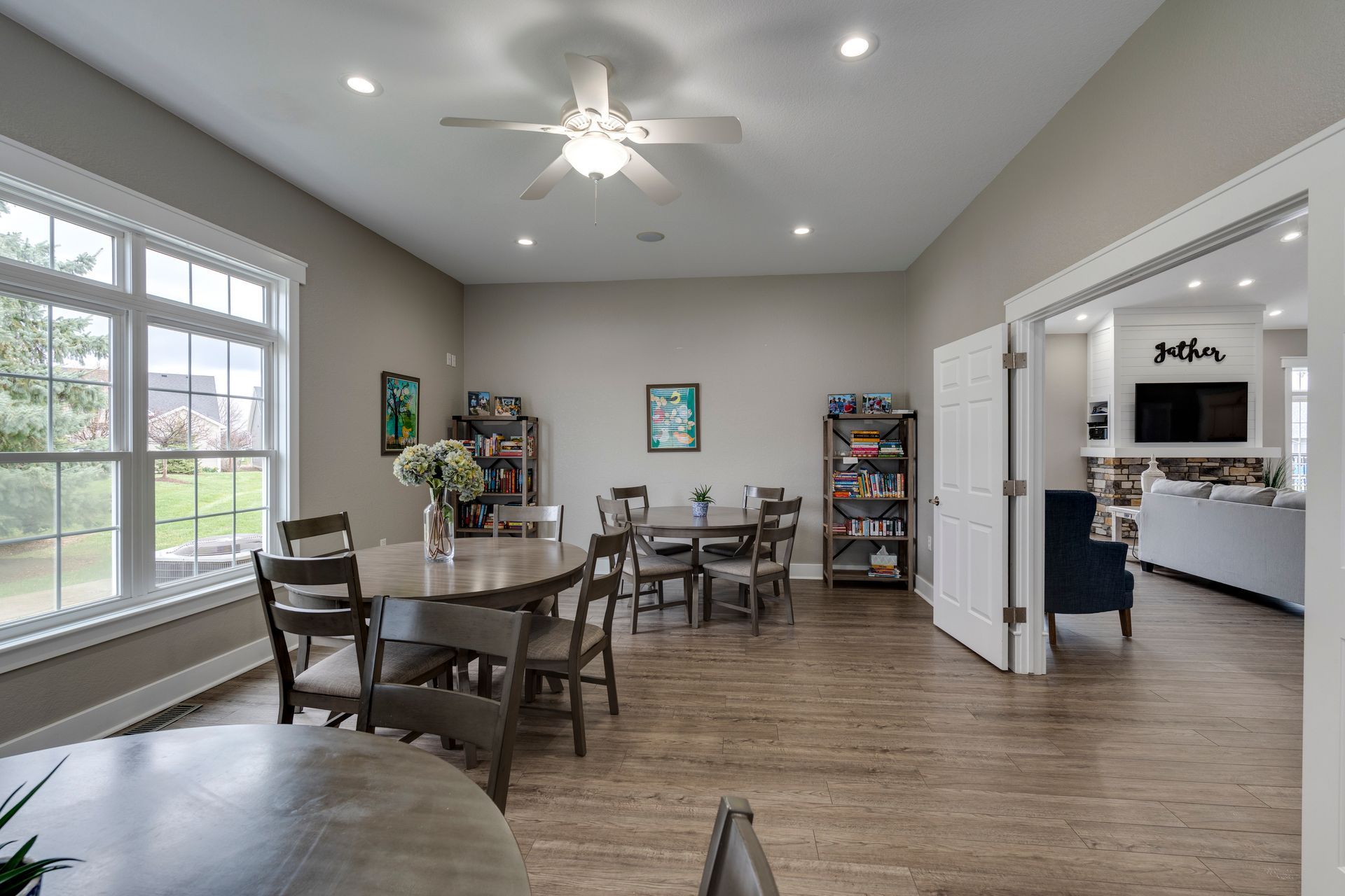 Spacious dining area with round tables, chairs, bookshelves, and large windows.  View into a living room with a fireplace.