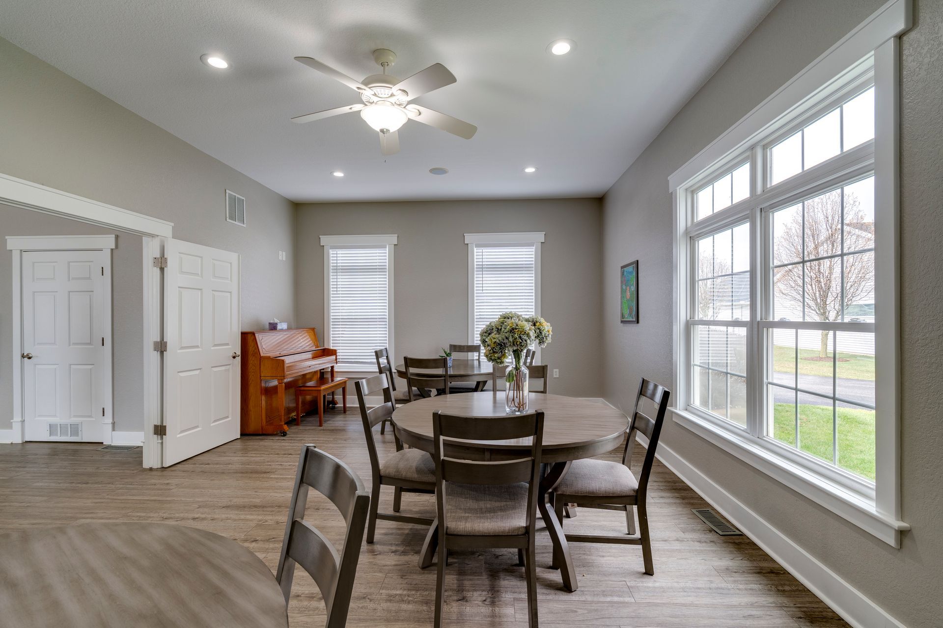 Large room with round tables, chairs, piano, and windows.