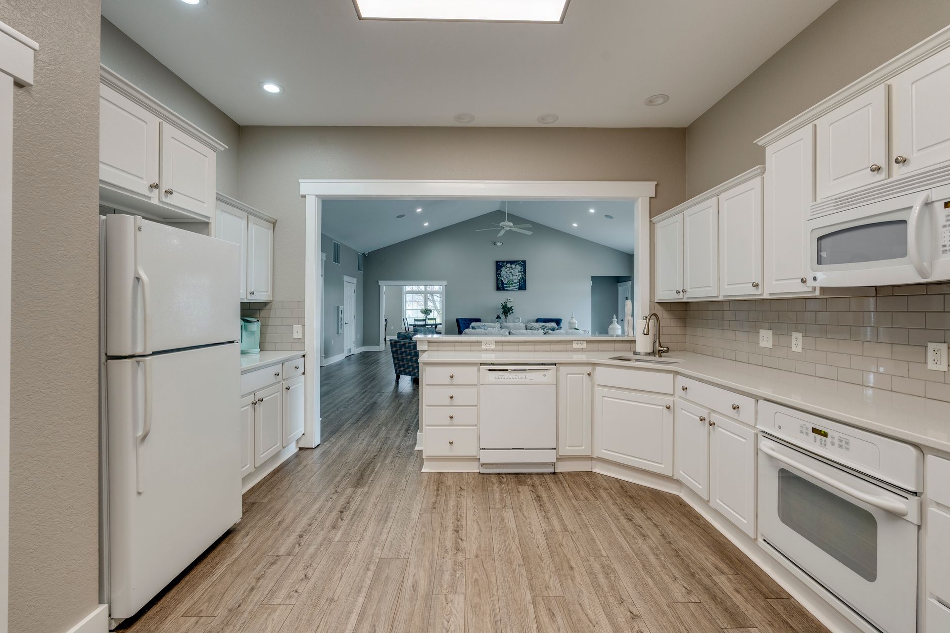 Bright white kitchen with a view into a living room, featuring light wood-look flooring and stainless steel appliances.