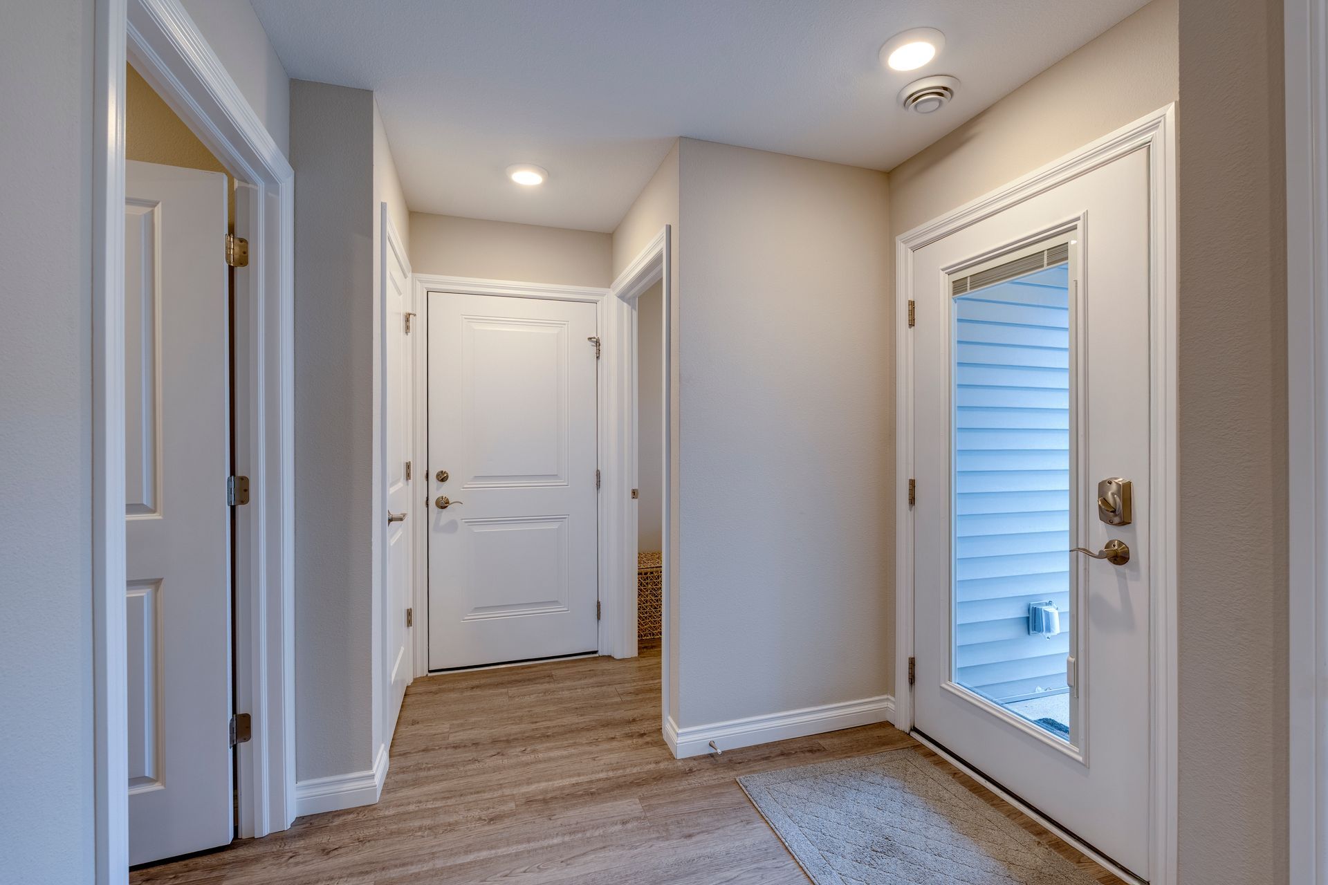 Hallway with three white doors, one with a window, beige walls, and wood-look flooring.