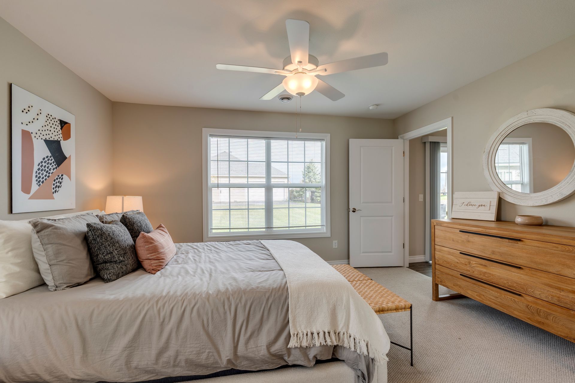 Bedroom with bed, dresser, window, artwork, and ceiling fan. Neutral color palette with light gray walls and beige carpet.