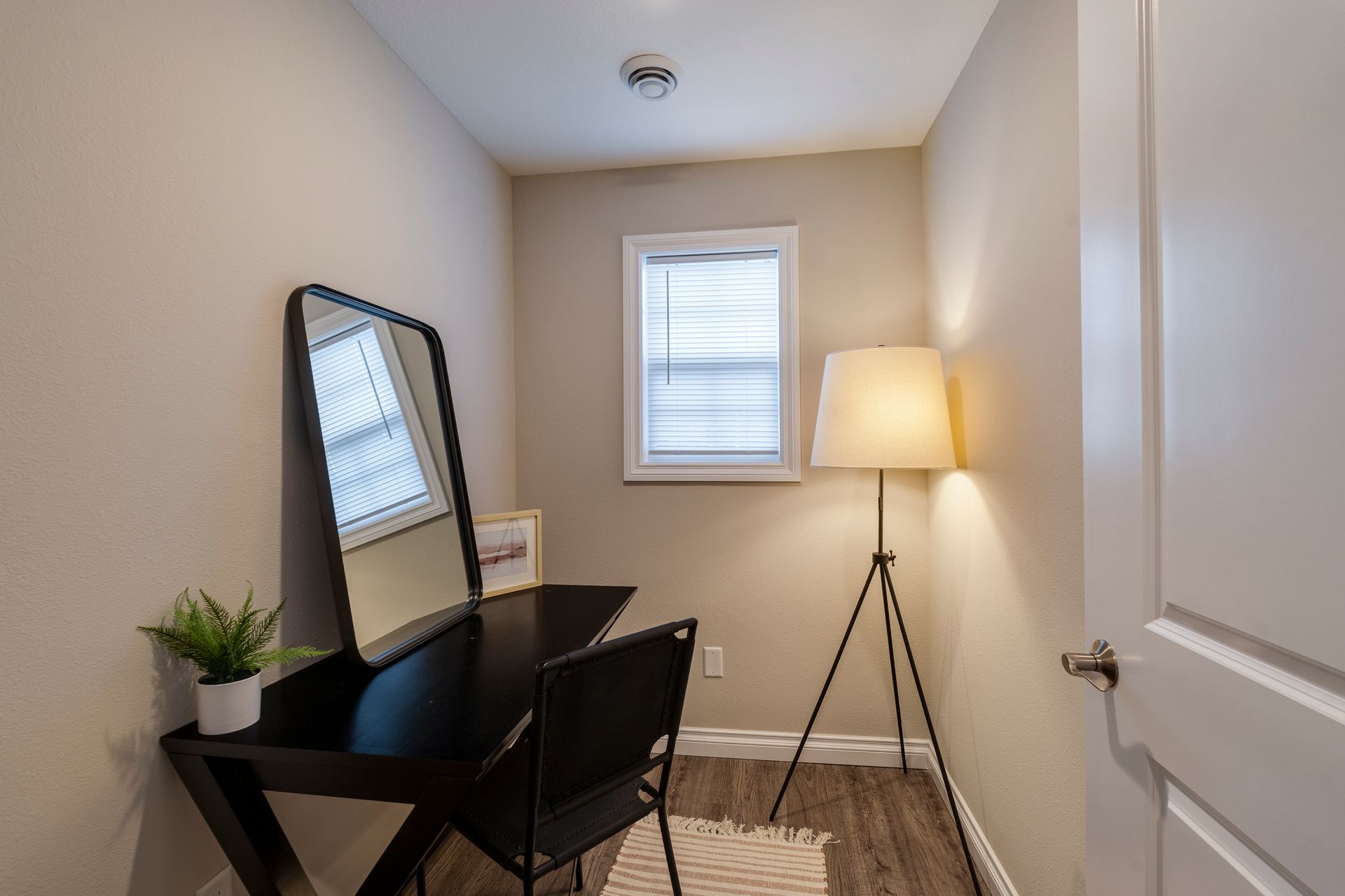 A makeup vanity with a black desk, mirror, and chair, next to a floor lamp, in a small room.