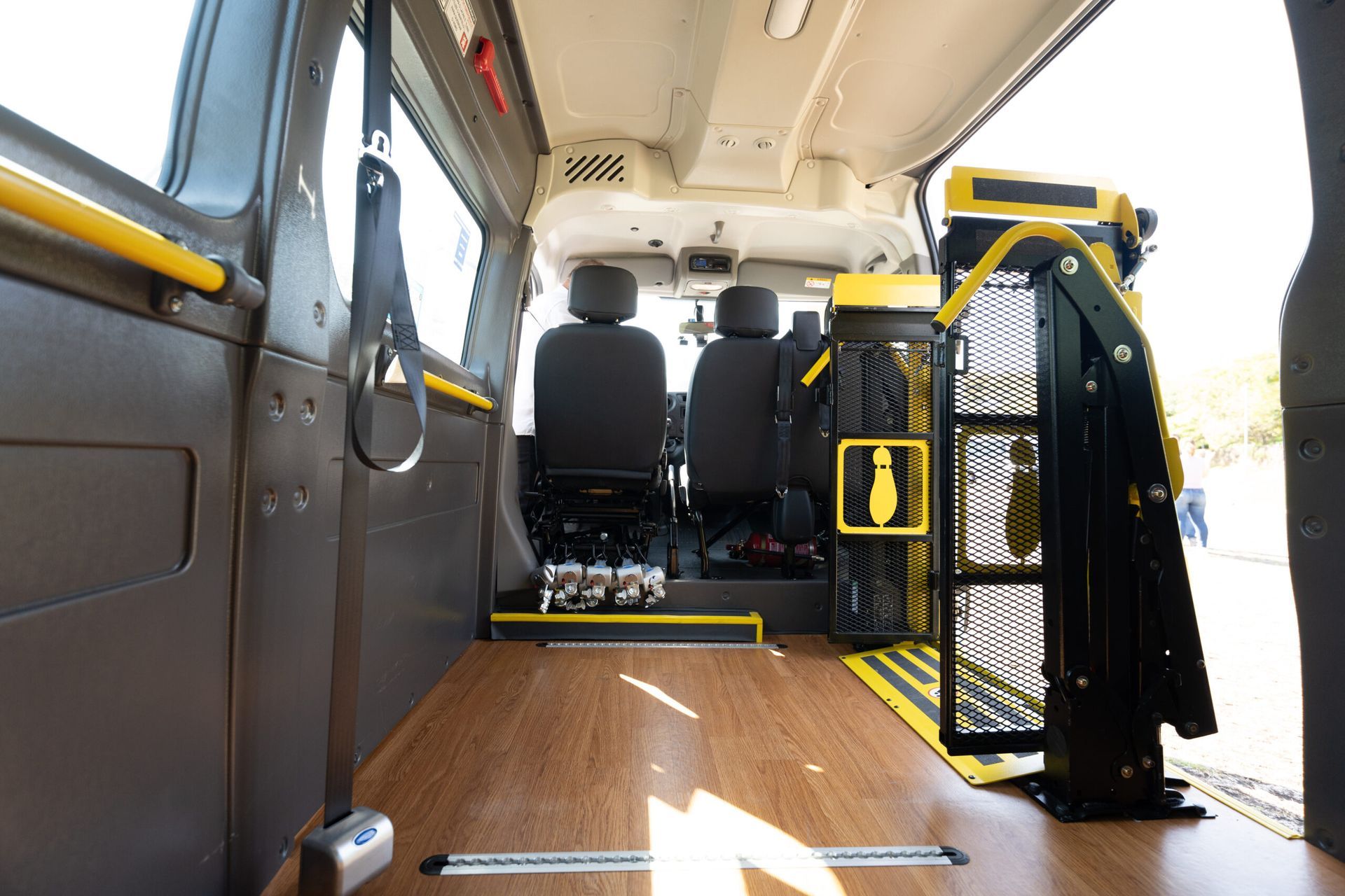 Interior de van adaptada com elevador para cadeiras de rodas, corrimãos amarelos e piso com padrão de madeira.