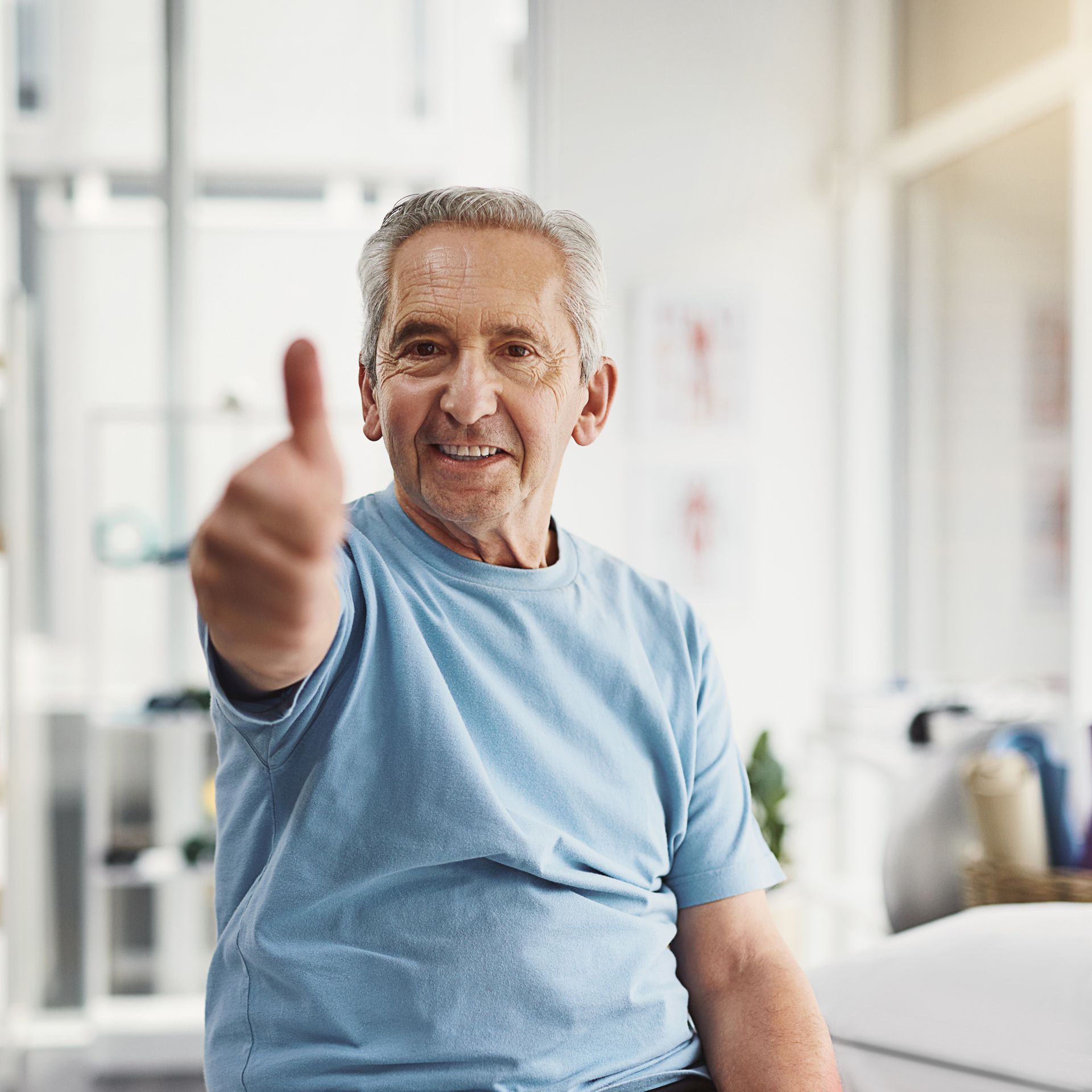 Homem sorridente de camisa azul faz sinal de positivo com o polegar, provavelmente em um ambiente médico ou de reabilitação.
