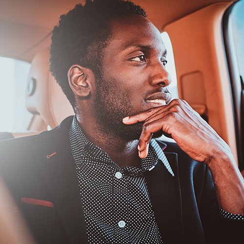 Homem de blazer preto e camisa estampada, com a mão no queixo, olhando pela janela de um carro.