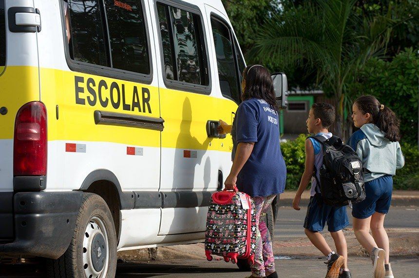 Crianças em idade escolar embarcando em uma van escolar amarela e branca; um adulto espera.