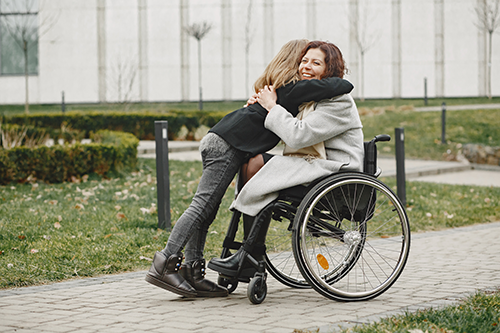 Uma mulher em cadeira de rodas abraça outra mulher em uma trilha pavimentada ao ar livre.