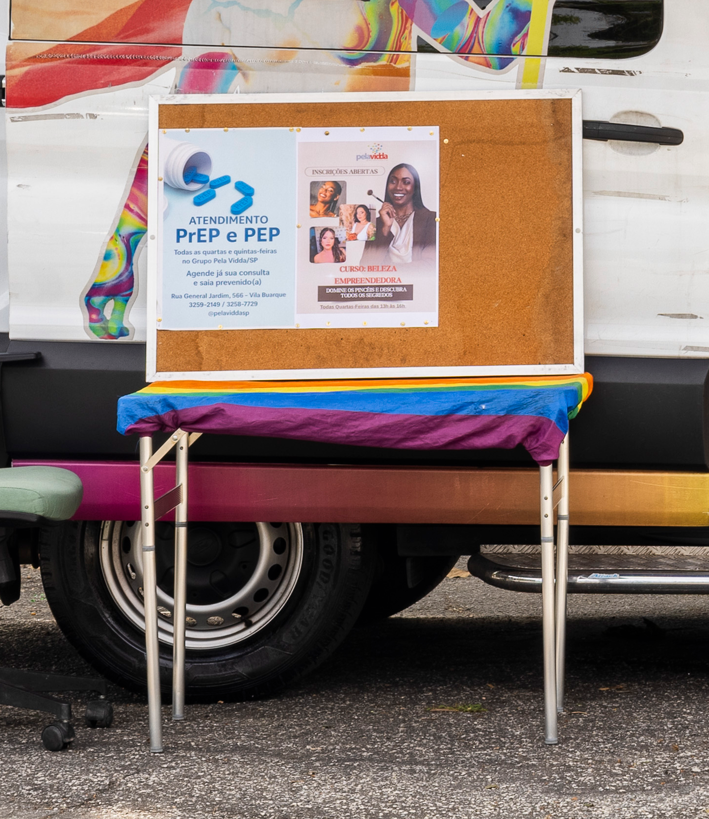 Quadro de cortiça com pôsteres sobre uma mesa em frente a uma van. A mesa tem uma toalha colorida.
