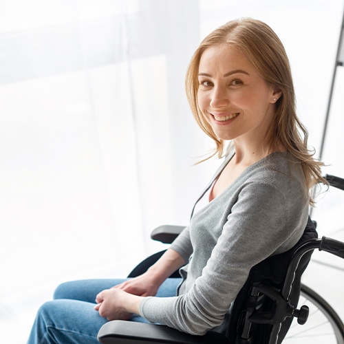 Mulher de longos cabelos loiros, sentada em cadeira de rodas, sorri perto de uma janela.
