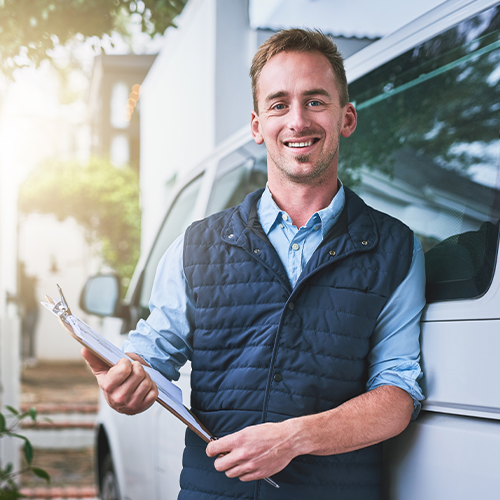 Homem de colete azul sorri, segurando uma prancheta, encostado em uma van. Ao ar livre, dia ensolarado.