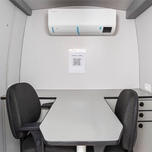 Interior de um pequeno escritório cinza com mesa, cadeiras, ar condicionado e um código QR.