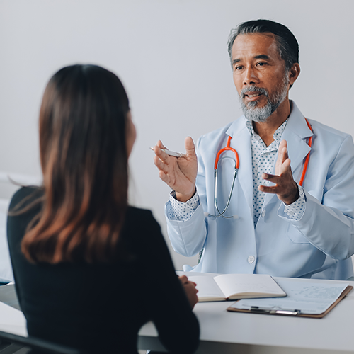 Médico de jaleco branco conversa com paciente sentado à mesa, com estetoscópio no pescoço.