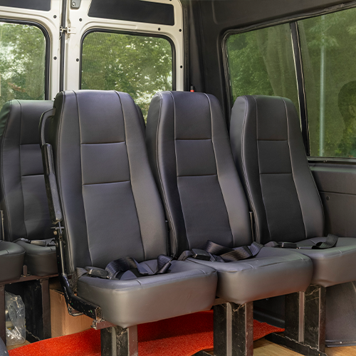 Interior de uma van com bancos e cintos de segurança pretos, contra um fundo branco e preto e piso vermelho.